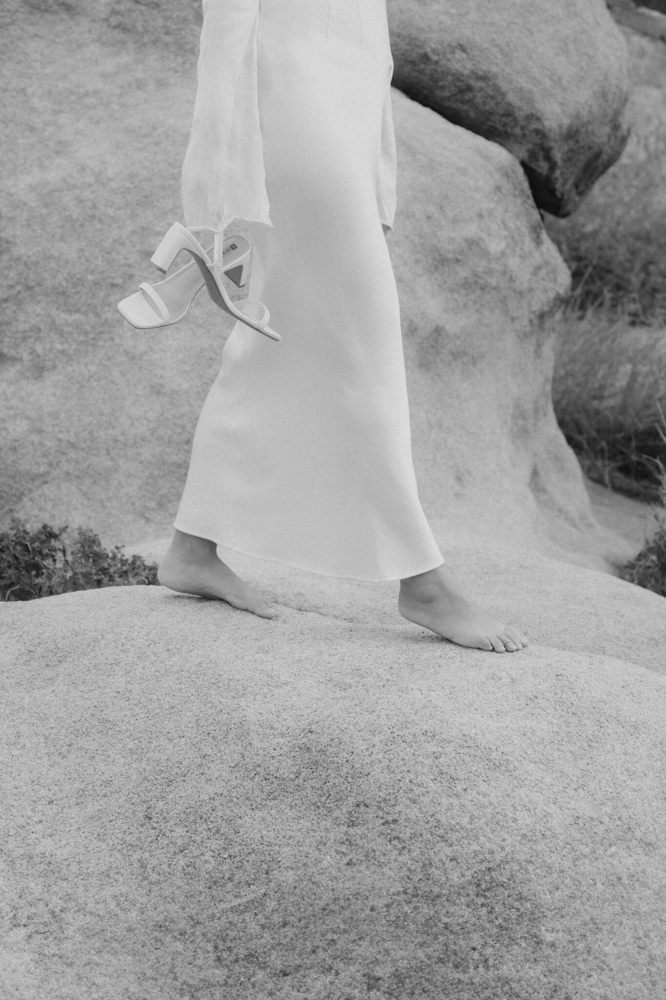 Barefoot bride walking on boulders, holding heels, minimalist black-and-white on their elopement in Europe