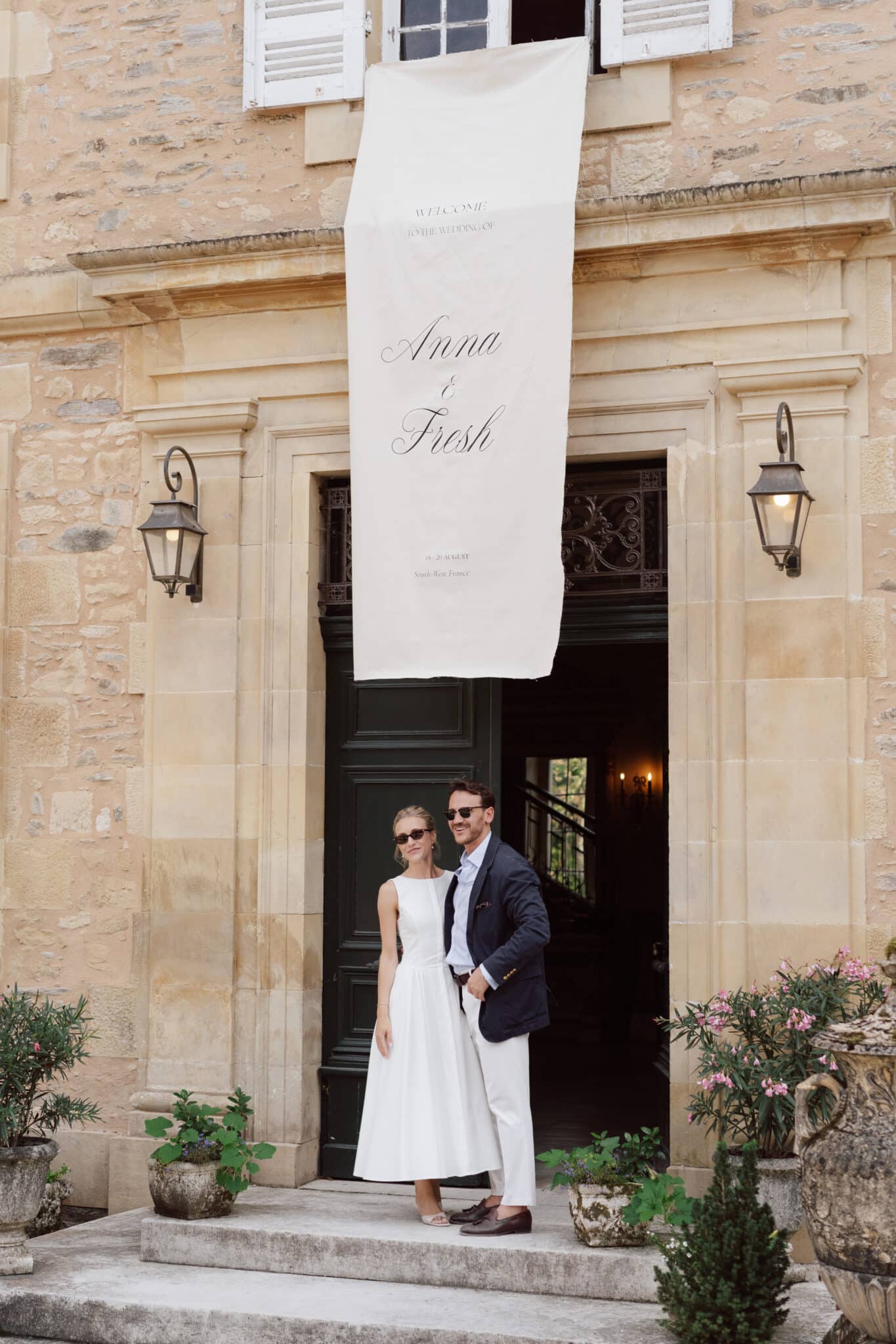Editorial portrait of bride and groom standing in the arched entrance of a French chateau, a personalized fabric banner overhead — destination wedding France